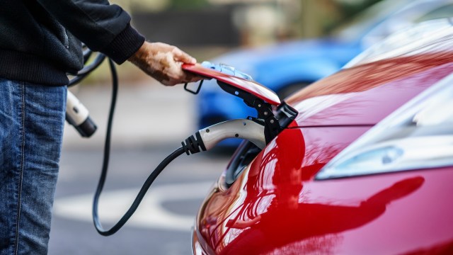 A person connects an electric vehicle to a charging station with a red car in the foreground.