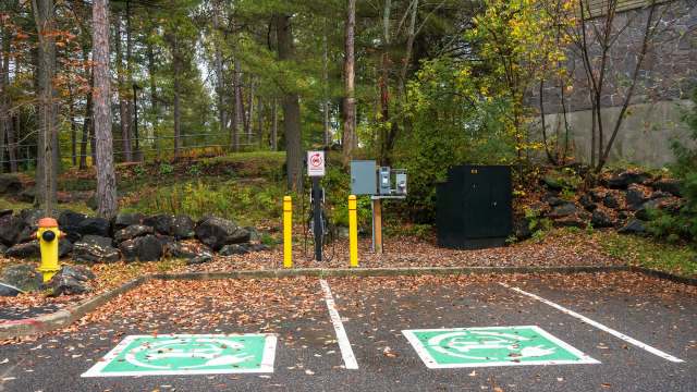 A parking lot with two spaces for electric vehicle charging, yellow bollards, and leaf-covered ground in a wooded area.