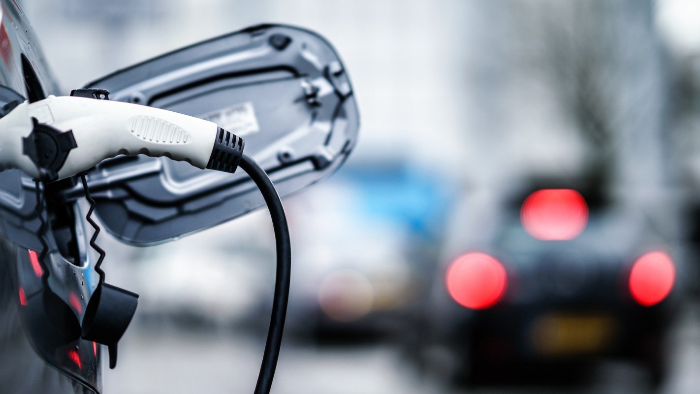 An electric car charging cable is plugged into a vehicle, with blurred traffic in the background.