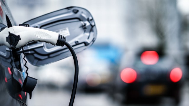 An electric car charging cable is plugged into a vehicle, with blurred traffic in the background.
