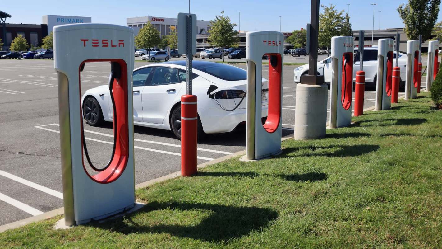A row of Tesla charging stations in a parking lot with a white Tesla vehicle parked nearby.