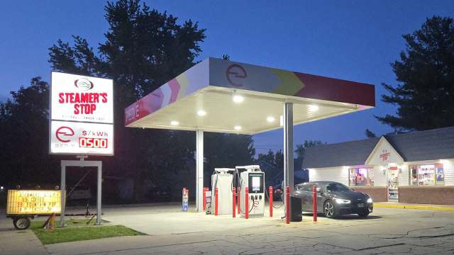 An illuminated service station named "Steamer's Stop" at dusk, with an EV parked and bright signs displaying prices.