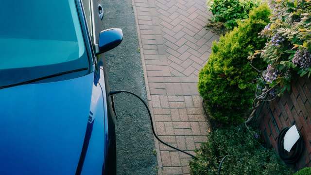 A blue electric car is charging with a cable beside a landscaped garden and brick pathway.