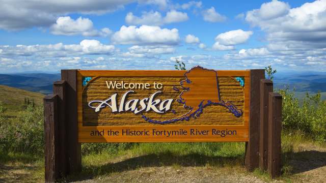 A wooden welcome sign for Alaska with scenic mountains and clouds in the background.
