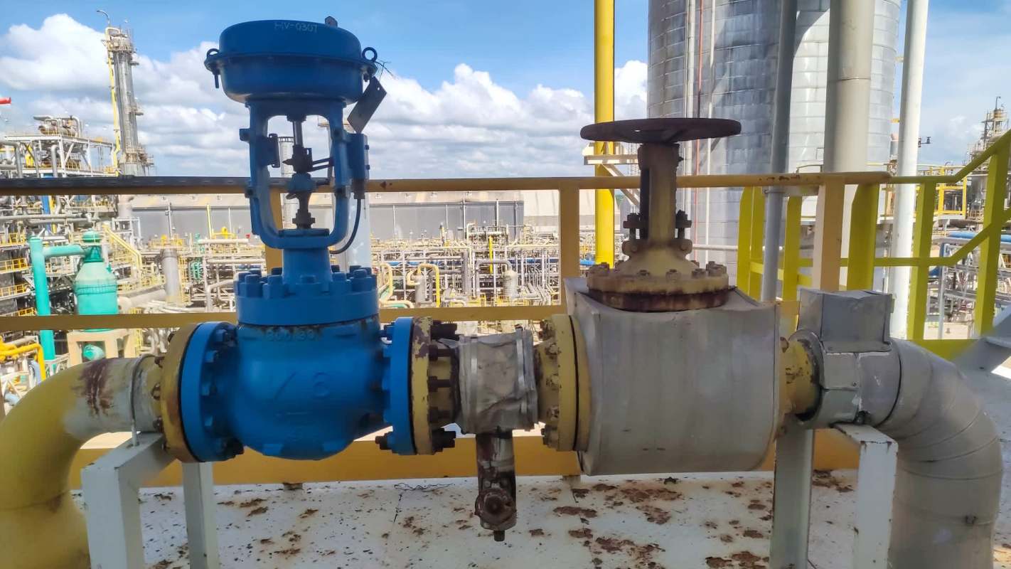 A blue industrial valve and a rusty pipe connected on a platform with a backdrop of industrial structures.