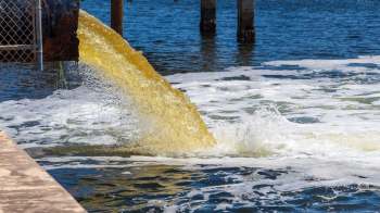 A pipe discharges murky water into a river, creating foam and ripples on the surface.