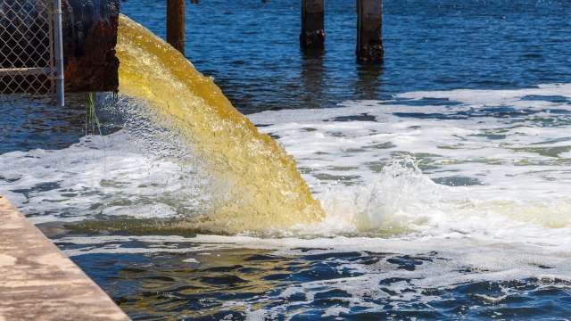 A pipe discharges murky water into a river, creating foam and ripples on the surface.