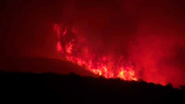 A large wildfire burns intensely against a dark landscape, glowing red amidst smoke.