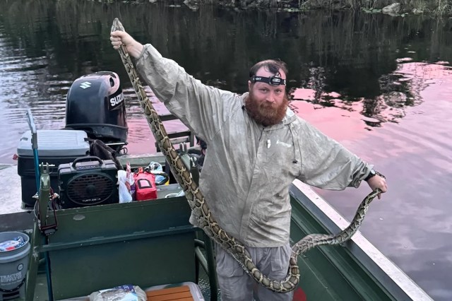 A man with a beard holds a large Burmese python while standing in a boat on a reflective body of water during sunset.