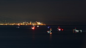 A city skyline illuminated at night with several boats lit up on the water in the foreground.