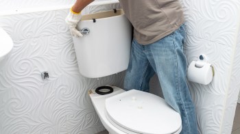 A person wearing gloves is installing a toilet tank in a bathroom with textured white walls.