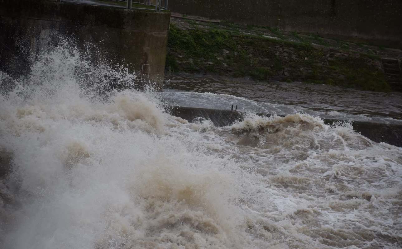 A turbulent river overflowing with muddy, churning water crashing against a concrete barrier.