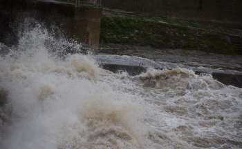 A turbulent river overflowing with muddy, churning water crashing against a concrete barrier.