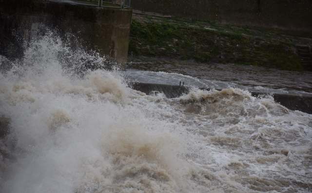 A turbulent river overflowing with muddy, churning water crashing against a concrete barrier.