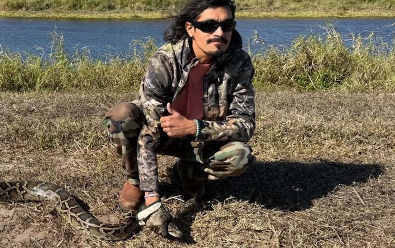 A person in camouflage clothing poses with a large snake by a riverbank under clear skies.