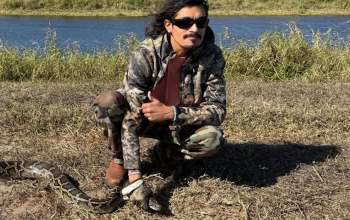 A person in camouflage clothing poses with a large snake by a riverbank under clear skies.