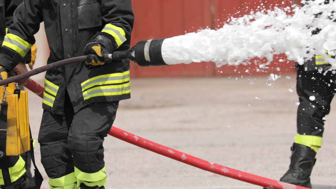 A firefighter sprays foam from a hose during a training exercise, wearing protective gear with reflective stripes.