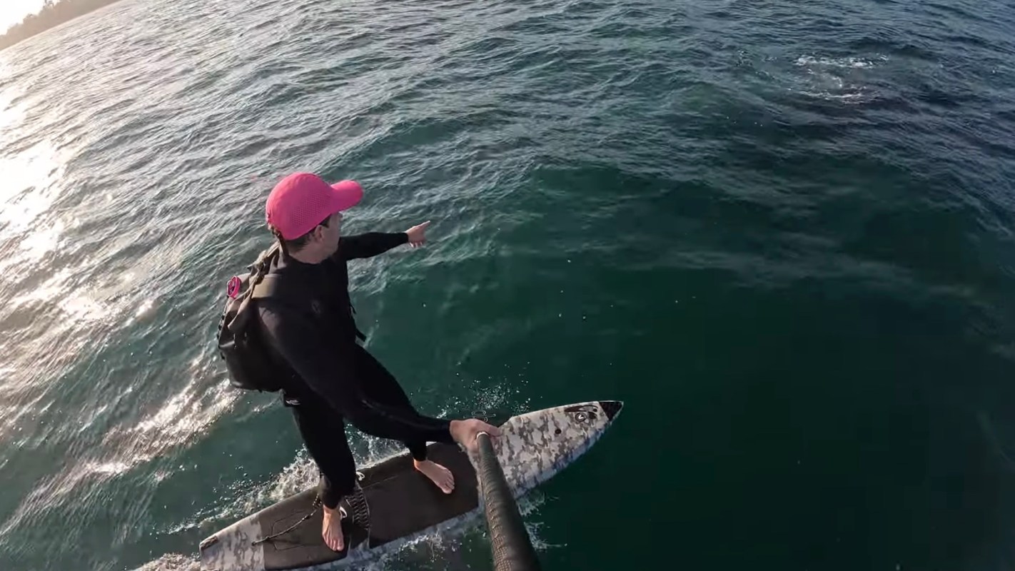 A person in a pink cap balances on a surfboard while pointing at something in the water.