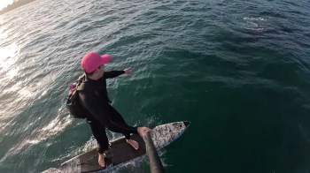 A person in a pink cap balances on a surfboard while pointing at something in the water.