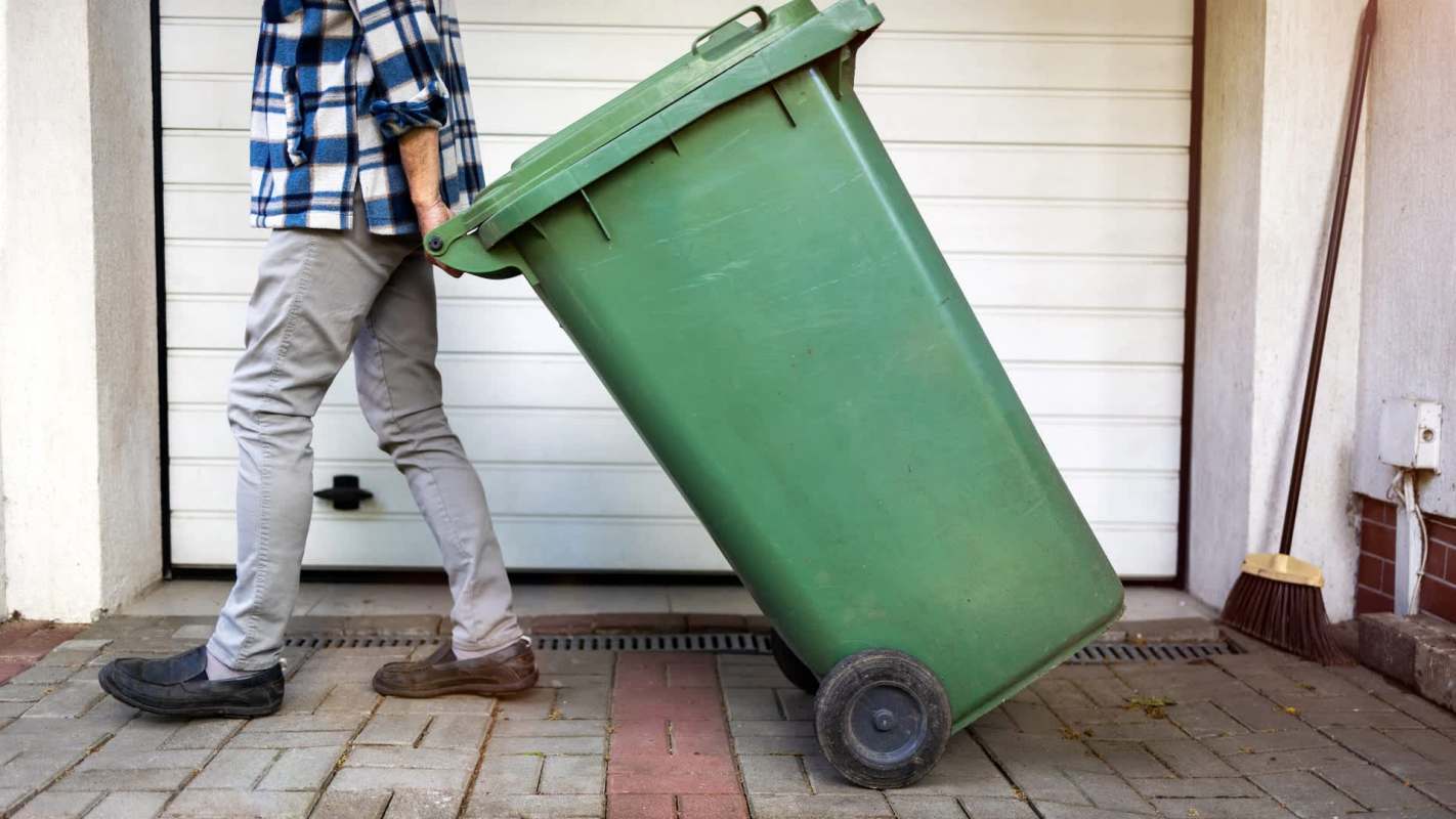 A person in a flannel shirt pushes a green wheelie bin along a paved path near a garage.