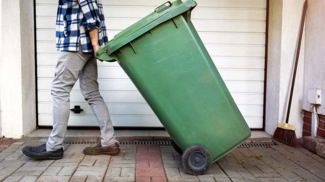 A person in a flannel shirt pushes a green wheelie bin along a paved path near a garage.