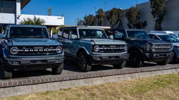Three Ford Bronco vehicles in various colors parked in front of a dealership under a clear blue sky.