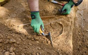 A gloved hand uses a trowel at an excavation site.
