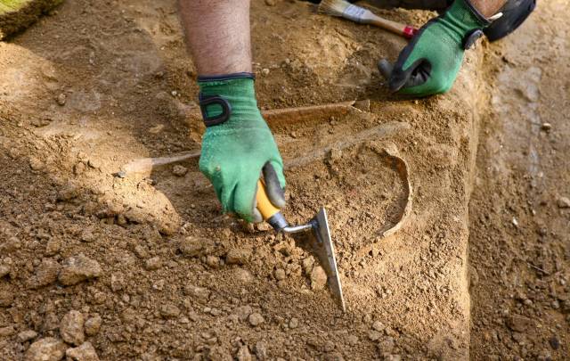 A gloved hand uses a trowel at an excavation site.