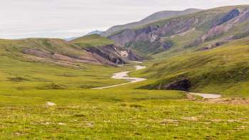 A winding river flows through a lush green valley surrounded by rugged mountains under a cloudy sky.