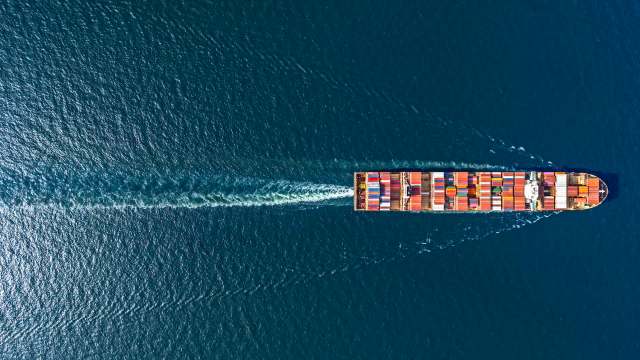 An aerial view of a cargo ship navigating through clear blue waters, leaving a wake behind.