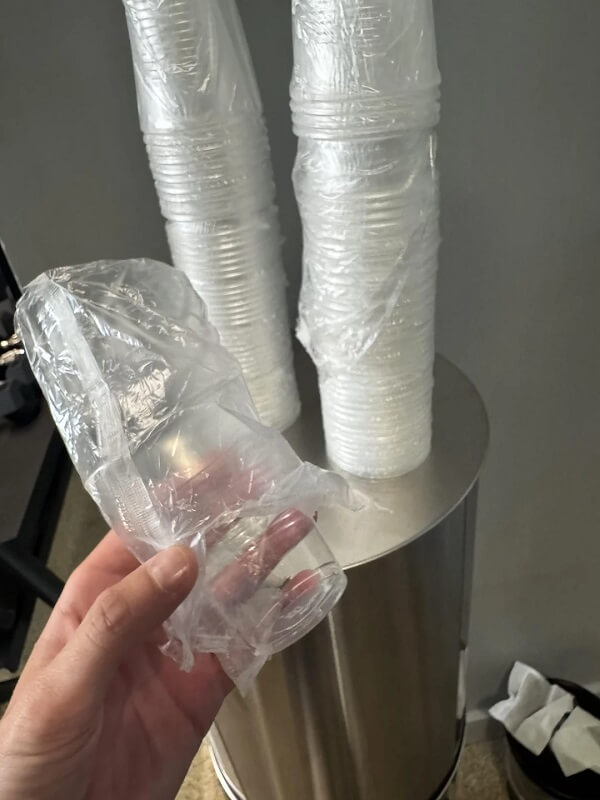 A hand holding a small clear plastic cup wrapped in crinkled film, with a metal trash can in the background.