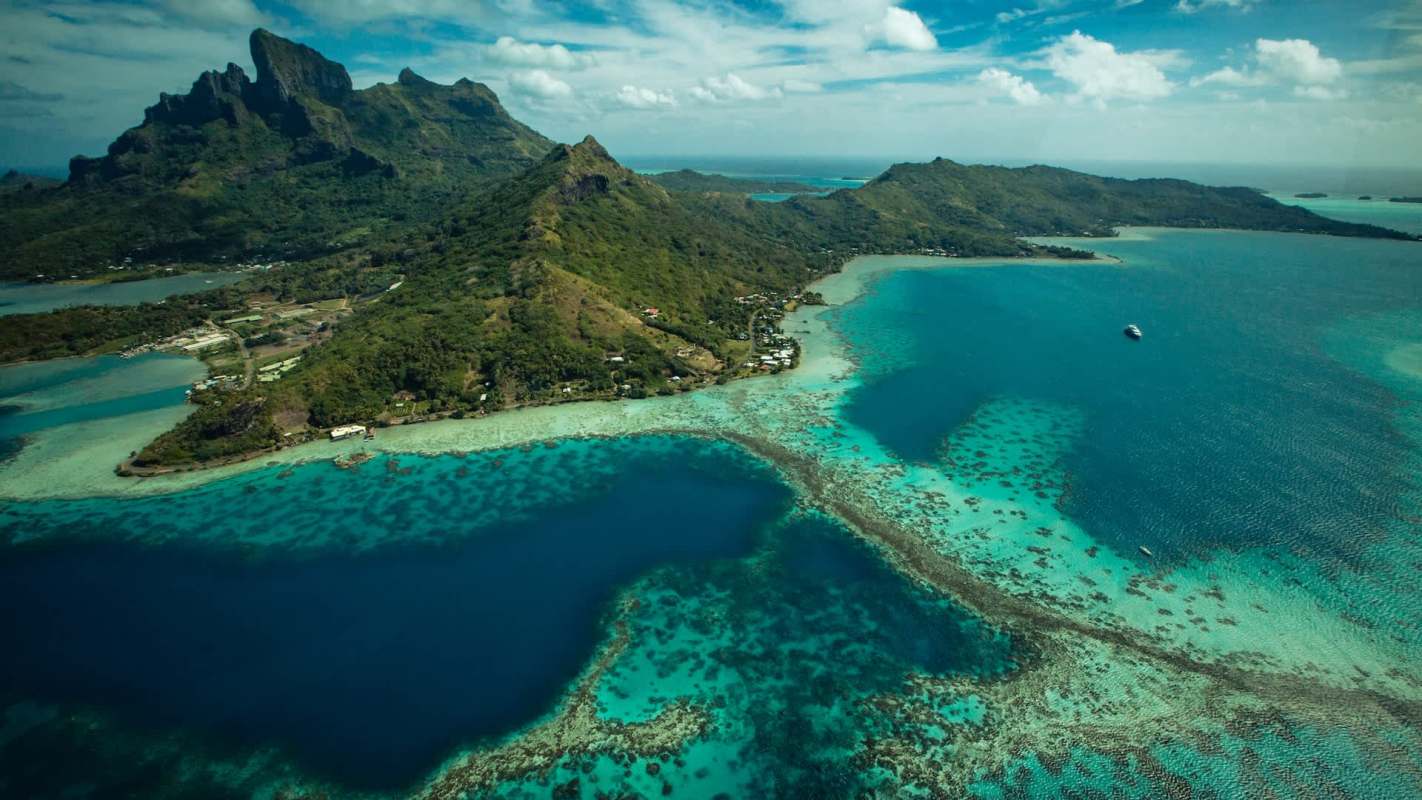 Aerial view of a lush green island with turquoise waters and coral reefs under a partly cloudy sky.