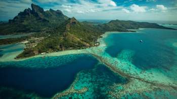 Aerial view of a lush green island with turquoise waters and coral reefs under a partly cloudy sky.