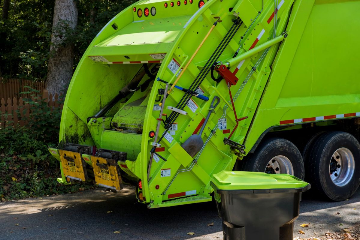A bright green garbage truck with an open rear compactor next to a bin on a residential street.