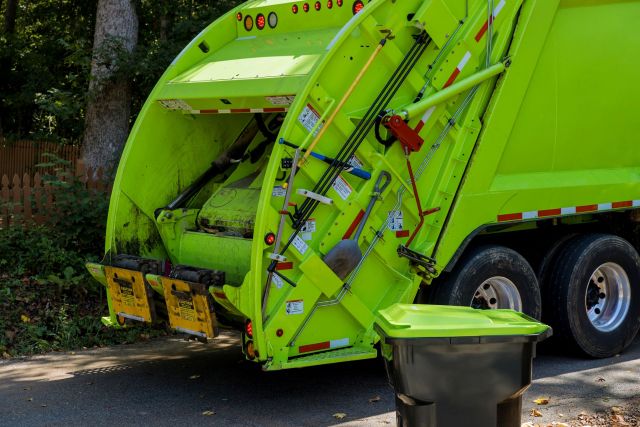 A bright green garbage truck with an open rear compactor next to a bin on a residential street.