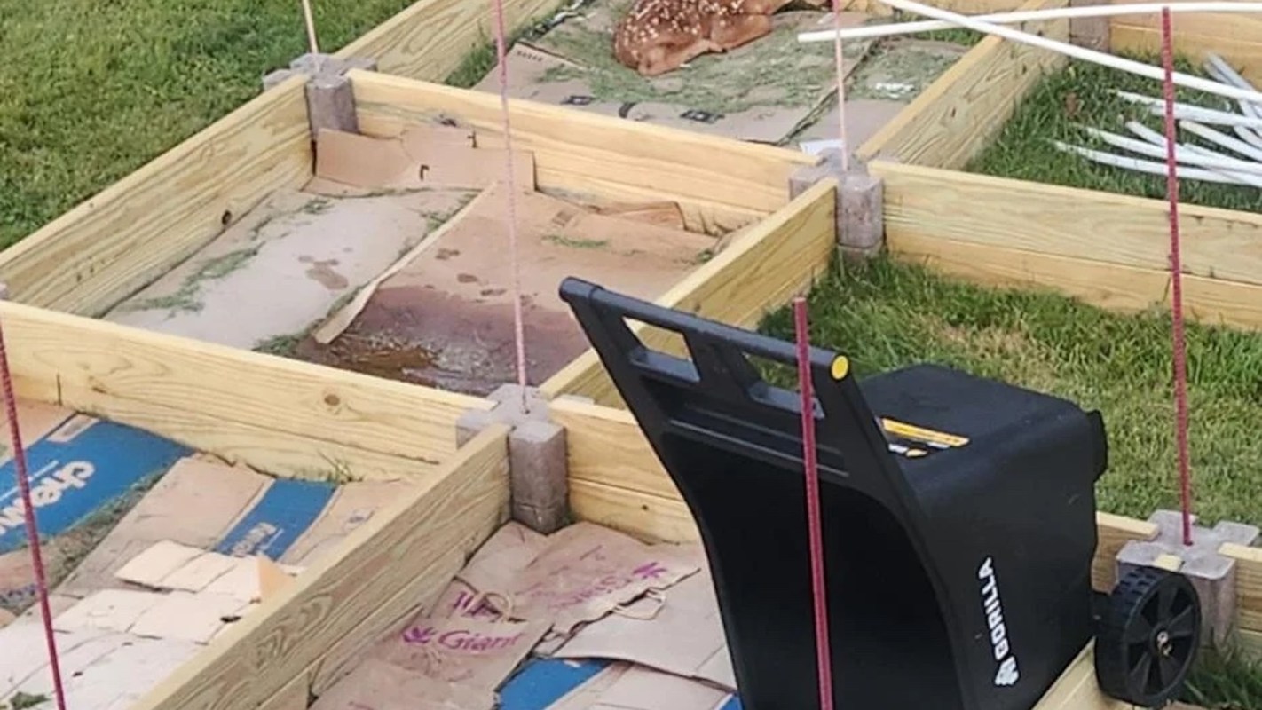 A black garden cart sits next to wooden planter boxes in a grassy area, with cardboard visible inside the boxes.