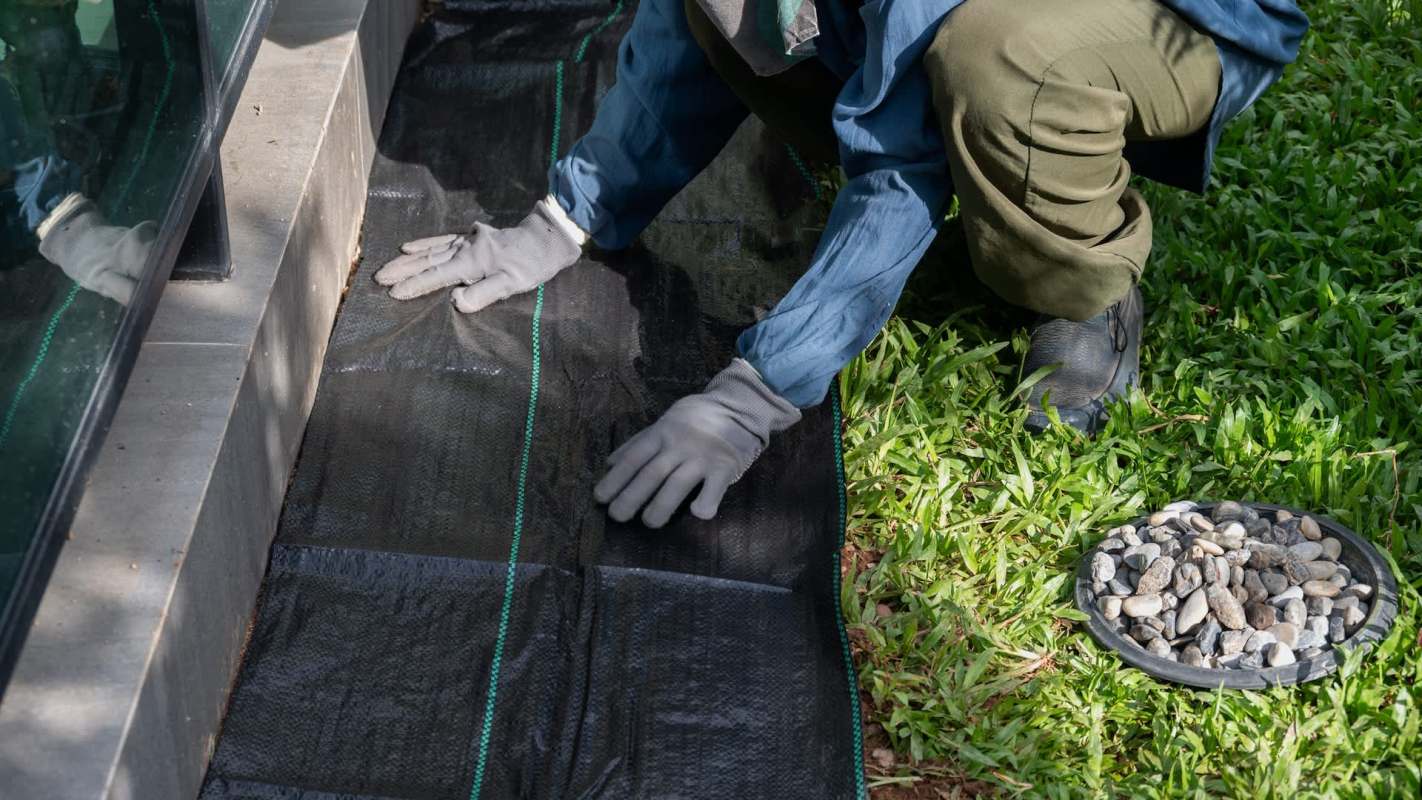A person in gloves lays down landscaping fabric next to a building, with a bowl of stones nearby on the grass.