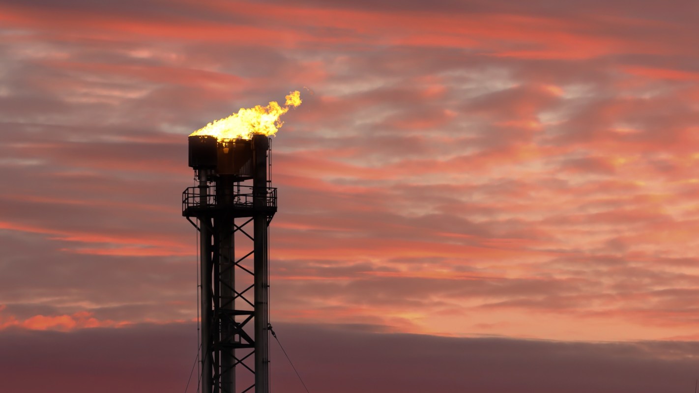 A gas flare burning brightly atop a tall industrial tower against a colorful sunset sky.