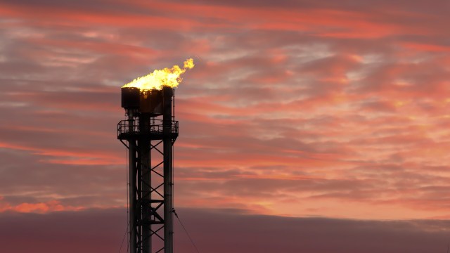 A gas flare burning brightly atop a tall industrial tower against a colorful sunset sky.
