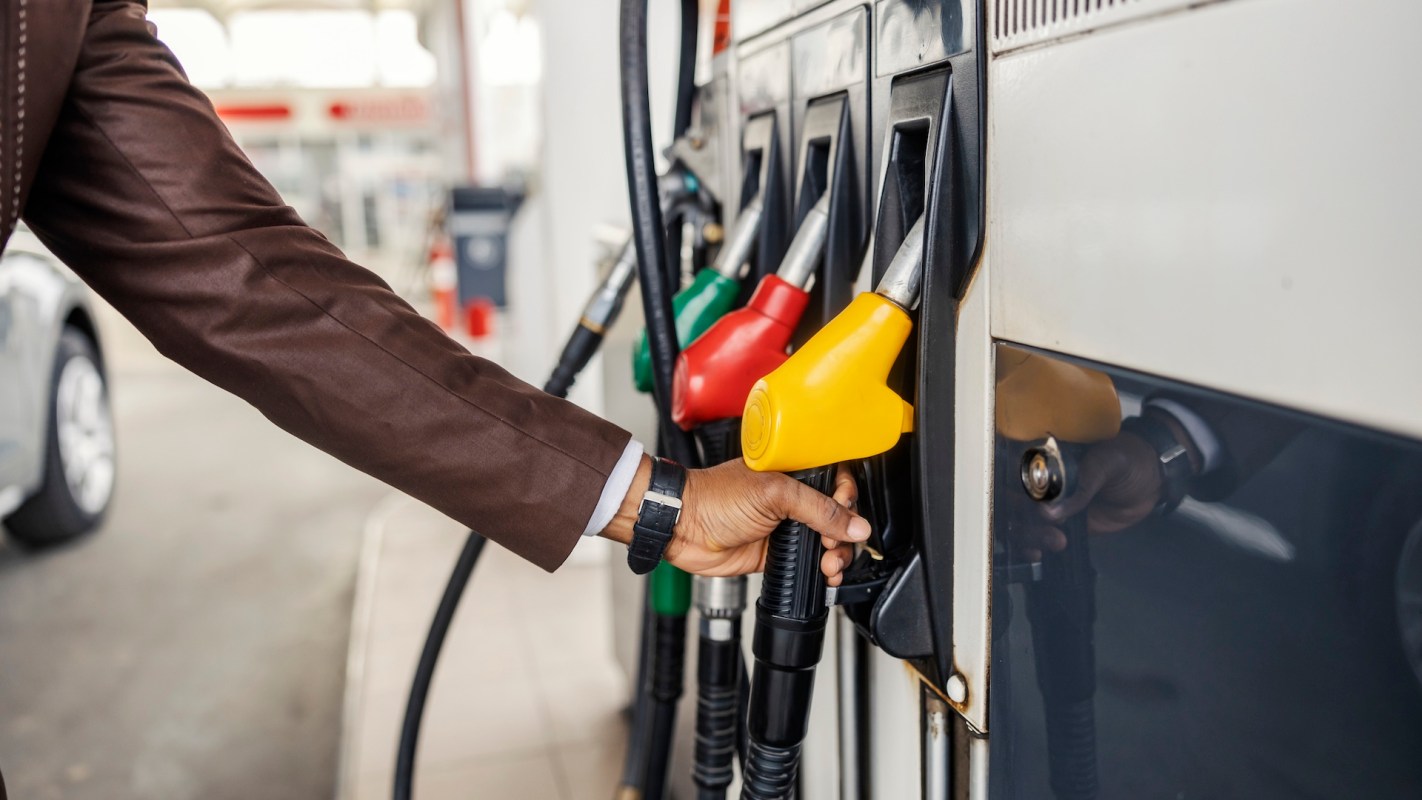 A person in a brown jacket holding a yellow fuel nozzle at a gas station pump.