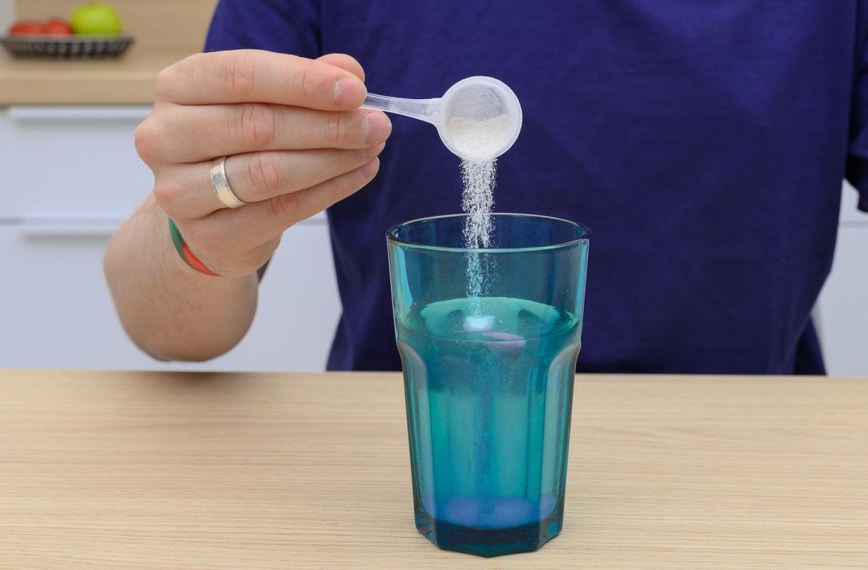 A person pouring a white powder from a measuring spoon into a turquoise glass of water on a wooden table.