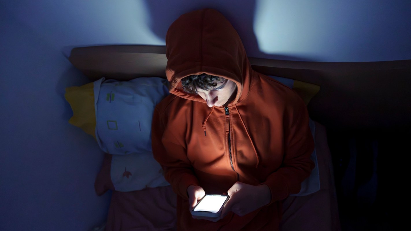 A person in an orange hoodie sits on a bed, using a smartphone that illuminates their face in a dark room.