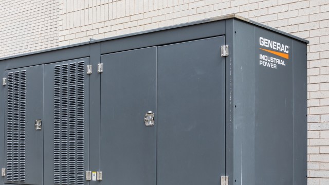 A large gray industrial power generator with ventilation panels against a brick wall.