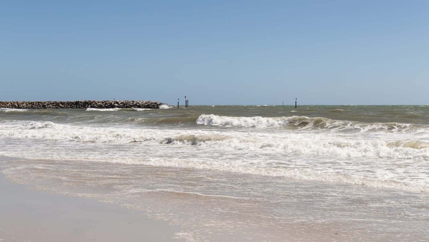 Waves crash onto a sandy beach with a rocky jetty and green markers in the background under a clear blue sky.
