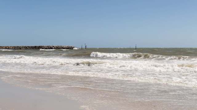 Waves crash onto a sandy beach with a rocky jetty and green markers in the background under a clear blue sky.