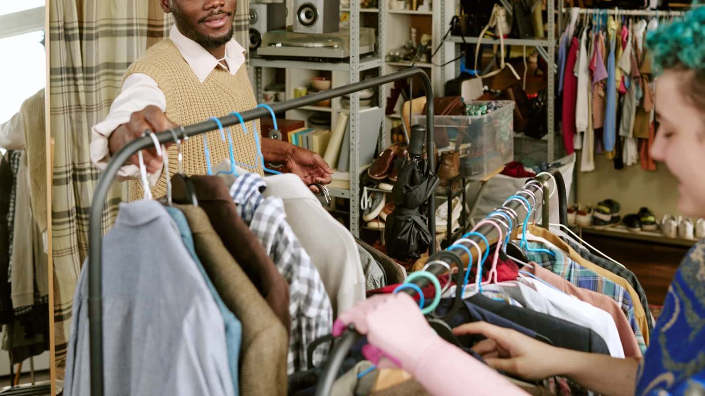 A person with short hair and a purple shirt examines clothes on a rack while another person smiles nearby.