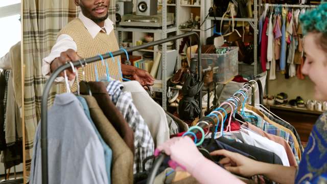 A person with short hair and a purple shirt examines clothes on a rack while another person smiles nearby.
