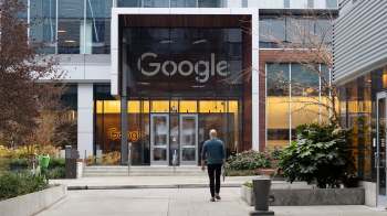 A person walks towards the Google building entrance, surrounded by greenery and modern architecture.