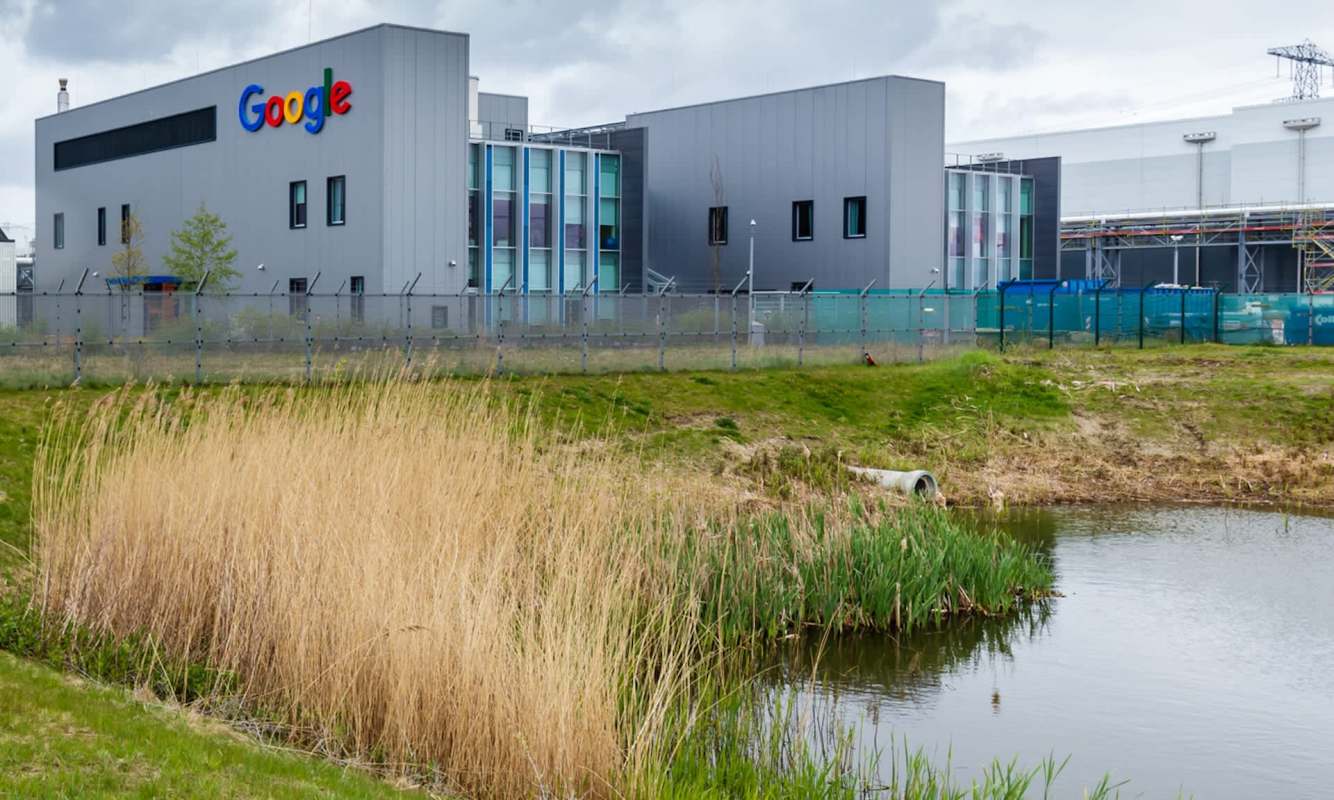 A Google data center beside a pond, surrounded by grass and reeds under a cloudy sky.