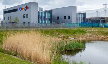 A Google data center beside a pond, surrounded by grass and reeds under a cloudy sky.